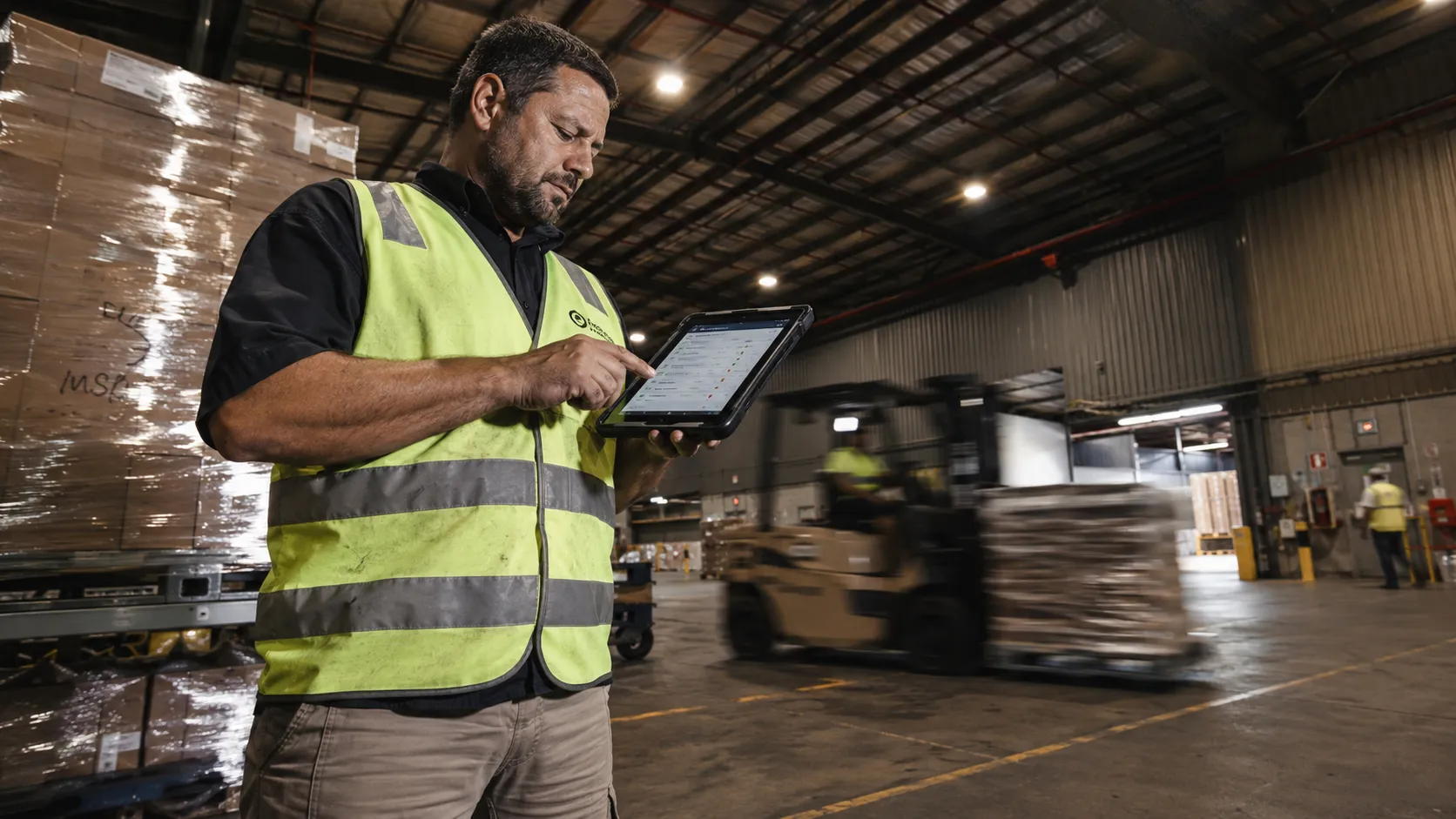 A warehouse worker in a hi-vis vest checking stock on a tablet, a forklift moving pallets behind him